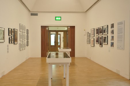 Interior of exhibition with works hung on walls and displayed in vitrines